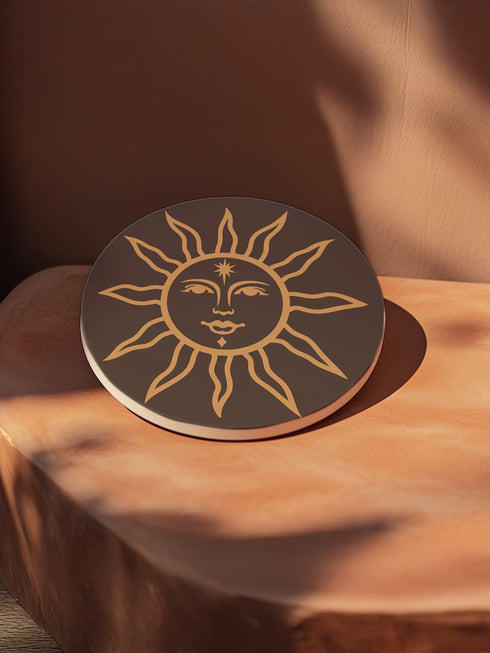 Round ceramic coaster featuring a celestial sun design in warm golden tones against a deep matte black background. The intricate illustration showcases a serene sun face with delicate detailing, surrounded by radiant, curved rays. The smooth, durable surface is complemented by a cork backing for grip and protection, making it both a functional and decorative piece for any space.